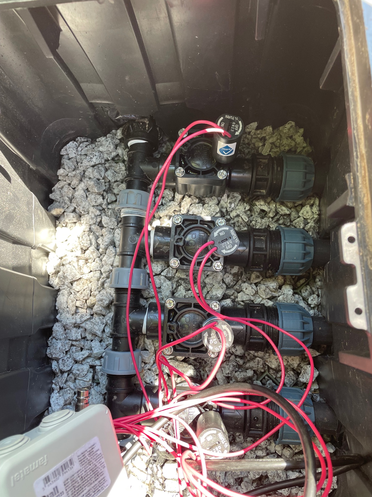 Skrzynka z zaworami i przewodami systemu nawadniania, wypełniona drobnym kamieniem, widok z góry.