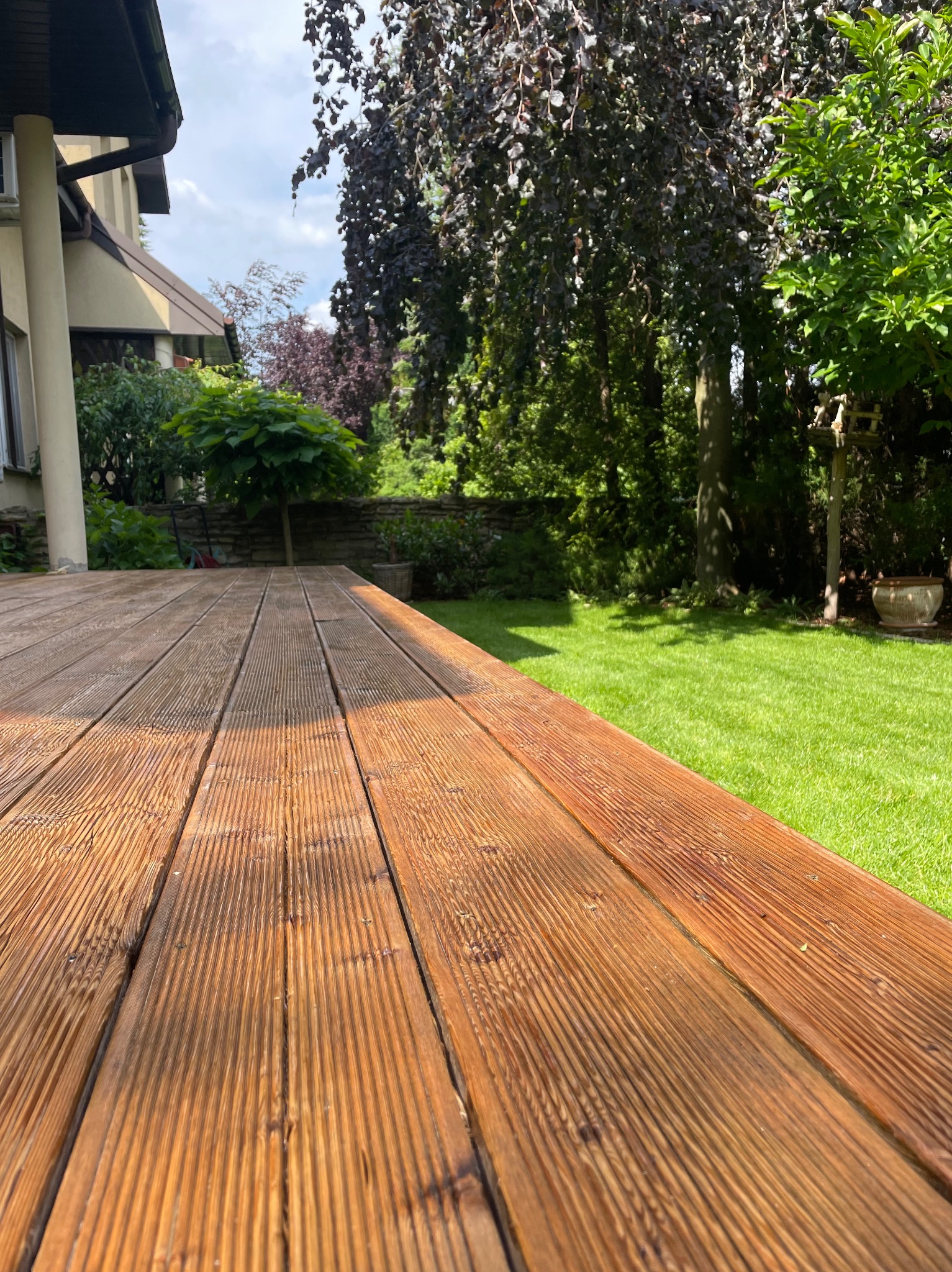 Drewniany taras z desek ryflowanych, prowadzący do zadbanego ogrodu z trawnikiem, drzewami i elementami architektury ogrodowej, widok z niskiej perspektywy.