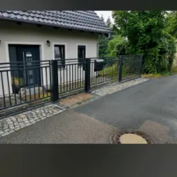 Nowoczesne czarne ogrodzenie metalowe z bramą i furtką, oddzielające posesję od ulicy z kostką brukową i studzienką kanalizacyjną, na tle domu z szarą elewacją i ciemną dachówką.