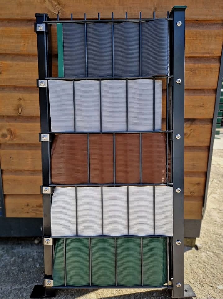 Ekspozycja paneli ogrodzeniowych w różnych kolorach (szary, biały, brązowy, zielony) na metalowym stojaku, na tle drewnianej elewacji.