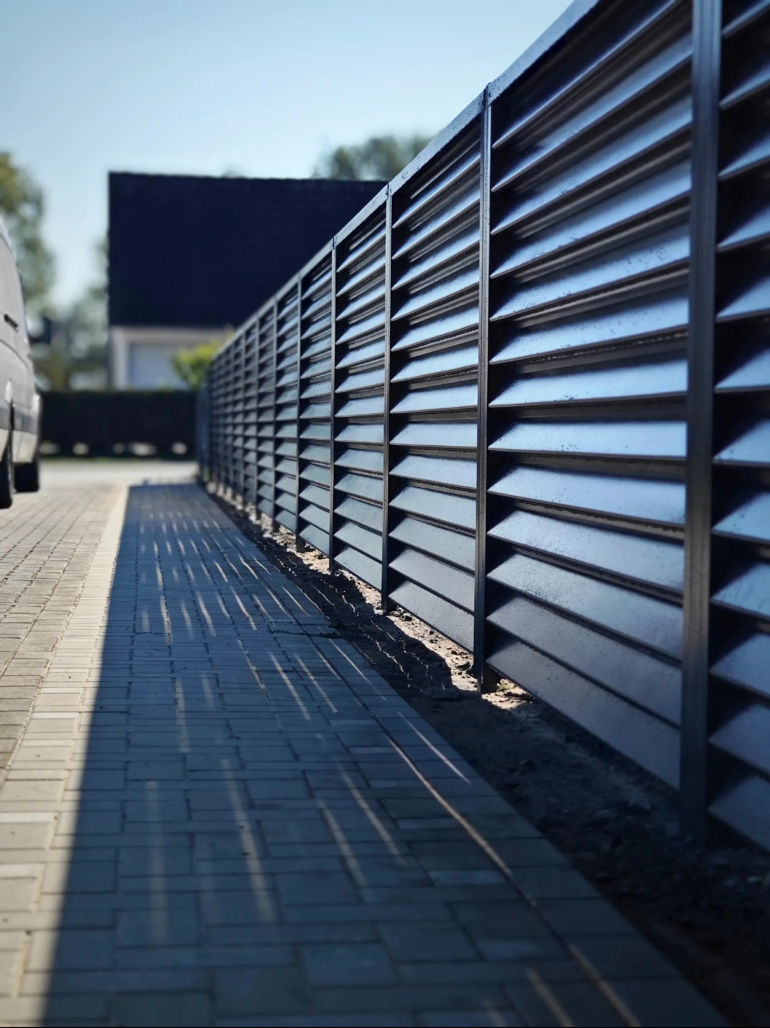 Nowoczesne, metalowe ogrodzenie lamelowe w kolorze grafitowym, rzucające cienie na brukowany chodnik, z fragmentem białego samochodu i domu w tle.