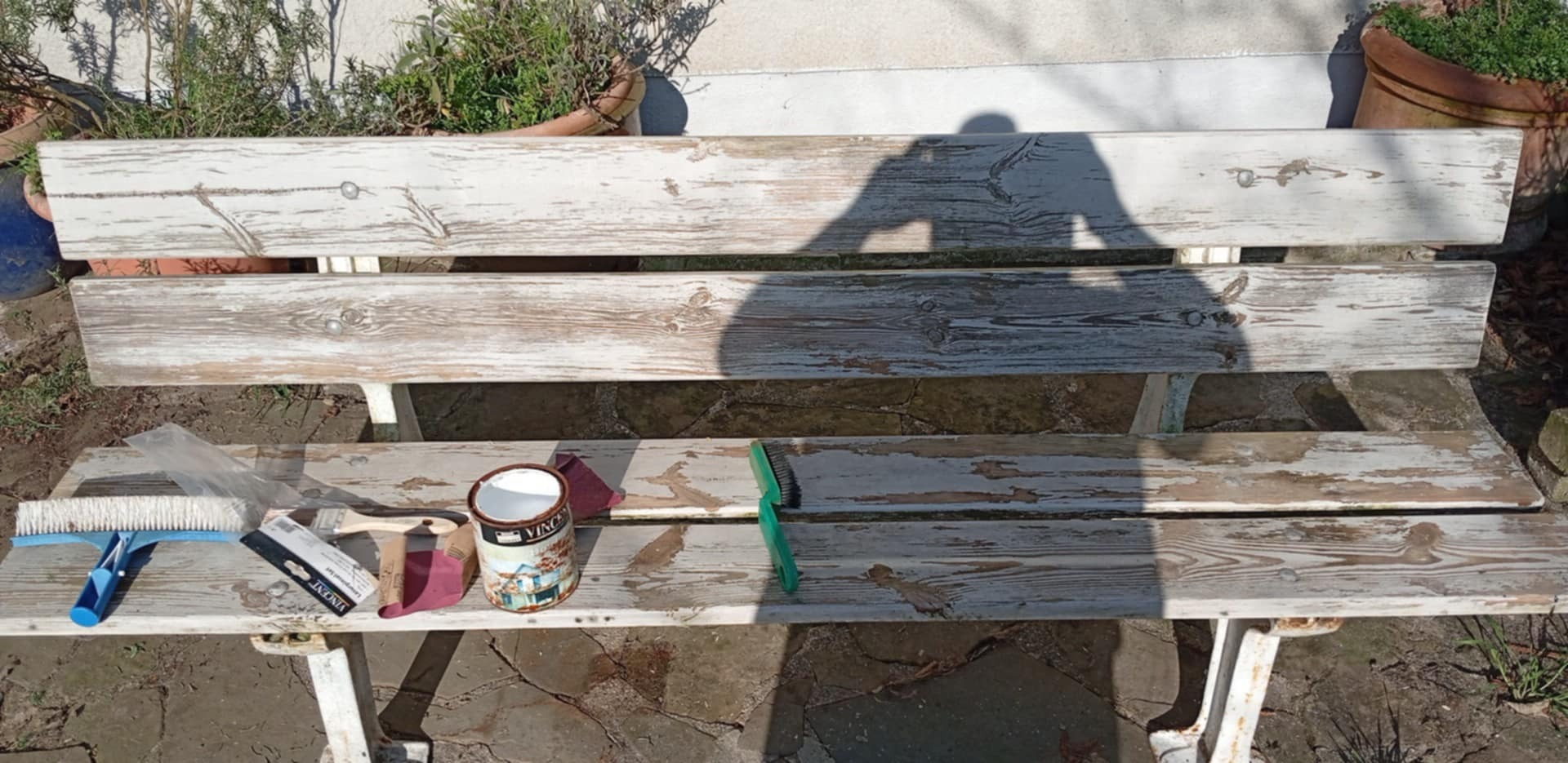 Renowacja ławki ogrodowej w toku: stara, drewniana ławka z resztkami białej farby, pędzle, papier ścierny i puszka farby Vincent na siedzisku w promieniach słońca.