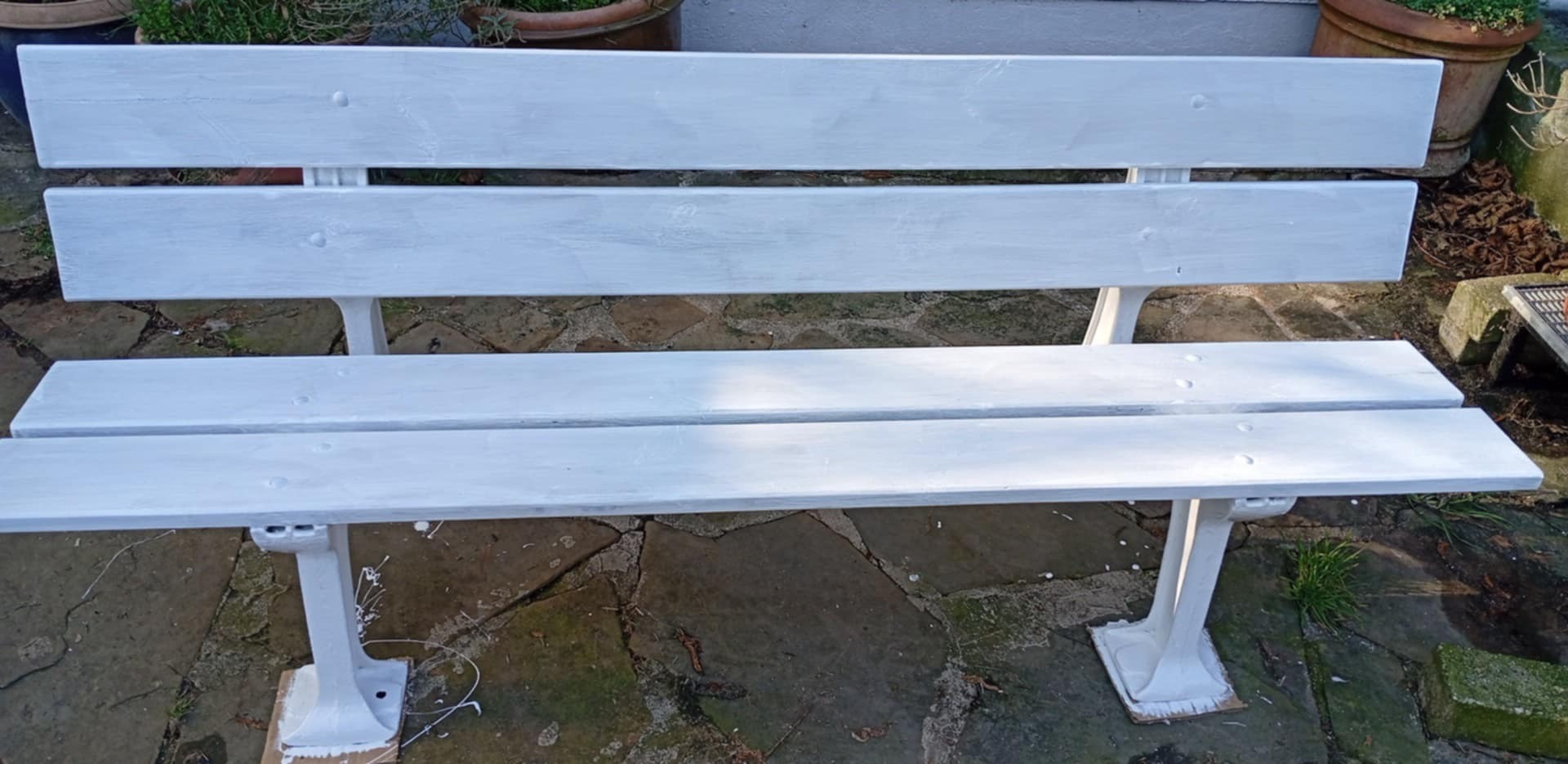 Odnowiona, biała ławka ogrodowa z drewnianym siedziskiem i metalowymi nogami, otoczona roślinnością i donicami. Świeżo pomalowana, widoczne drobne niedociągnięcia.