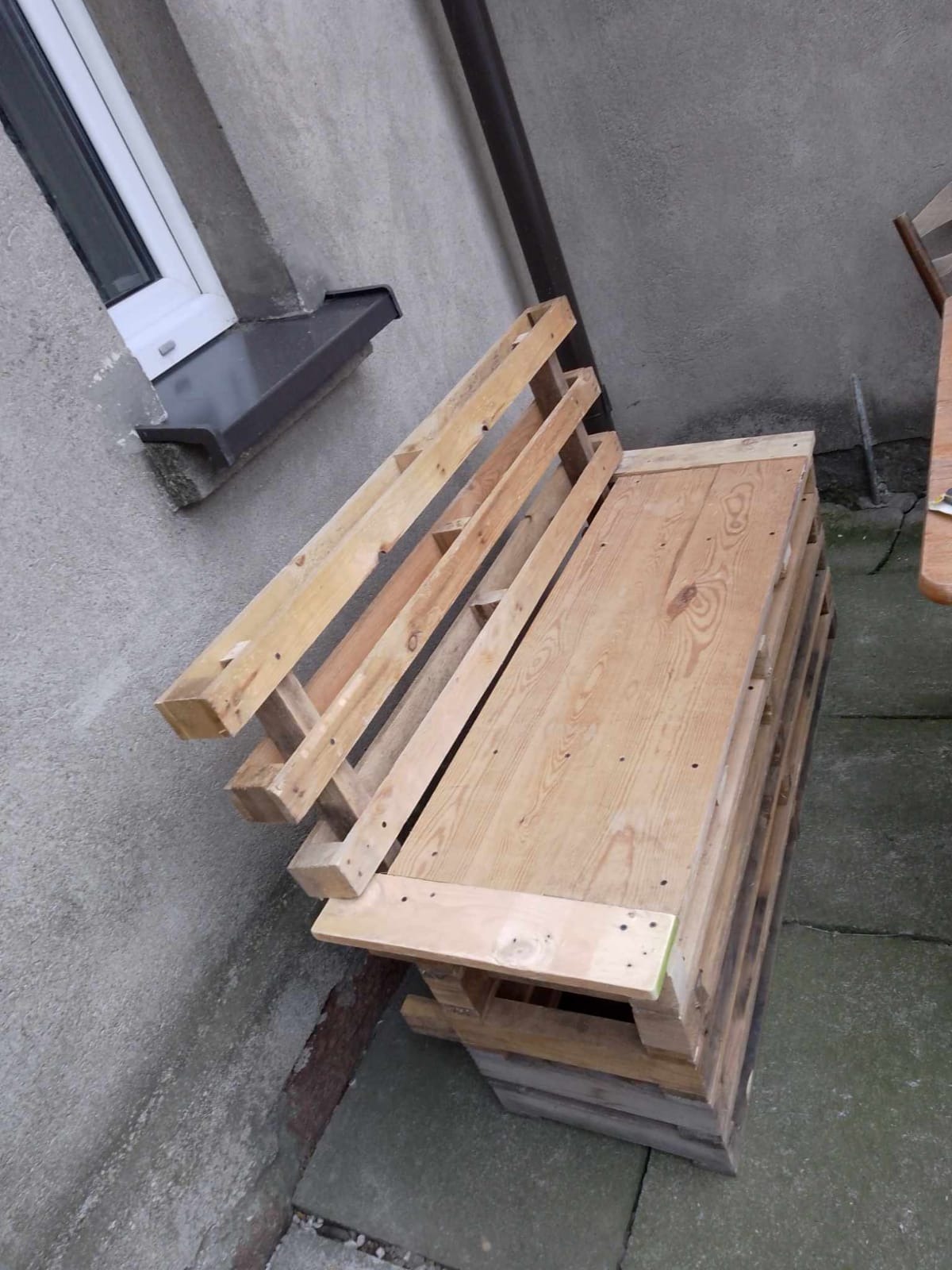 Niezwykła konstrukcja z drewnianych palet przy szarej ścianie, prawdopodobnie mebel ogrodowy w trakcie tworzenia. Widoczne elementy stolarskie.