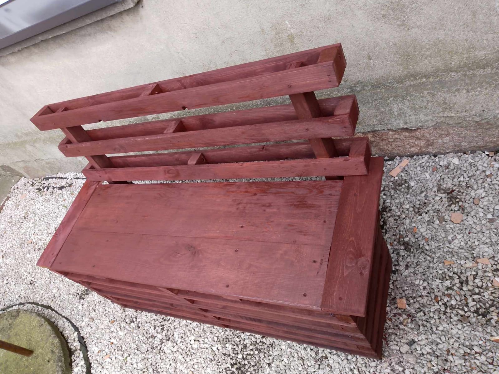 Drewniana konstrukcja w kolorze brązowym, przypominająca skrzynię z przegródkami na rośliny, ustawiona na żwirowym podłożu przy ścianie.