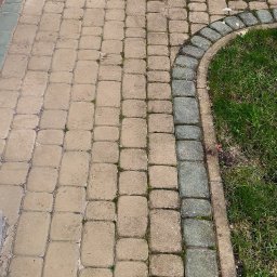 G&G SHOP MARZENA GRYGORUK - Częściowo wyczyszczona, beżowa kostka brukowa przed wejściem do domu, z widoczną różnicą w kolorze i czystości między obszarami po i przed czyszczeniem.  Widoczne zielone obrzeże z trawy po prawej...