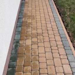 G&G SHOP MARZENA GRYGORUK - Świeżo ułożona, dwukolorowa kostka brukowa przylegająca do ściany budynku z widocznym odpływem liniowym, fragment kostki jest jeszcze mokry.