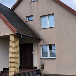 G&G SHOP MARZENA GRYGORUK - Dwukondygnacyjny dom z beżową elewacją i ciemnym dachem. Widoczne ślady zabrudzeń na elewacji, szczególnie pod oknami. Wejście z kolumną z żółtej cegły.