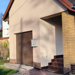 G&G SHOP MARZENA GRYGORUK - Dom z beżową elewacją, brązowym dachem i garażem. Widoczne schody z cegły klinkierowej i trawnik. Elementy instalacji na elewacji. Słoneczny dzień, błękitne niebo.