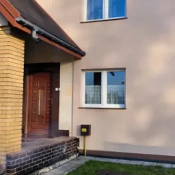 Jasna elewacja dwupiętrowego domu z brązowym dachem i drewnianymi drzwiami wejściowymi. Widoczne dwa okna na piętrze i jedno na parterze.