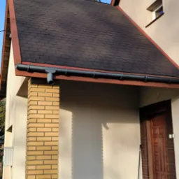 Dom z dachem pokrytym gontem bitumicznym w odcieniach brązu. Widoczna elewacja w jasnym kolorze, ceglana kolumna i drewniane drzwi wejściowe. Na dachu rośnie małe drzewko.