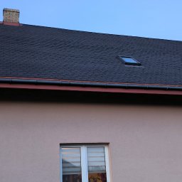 G&G SHOP MARZENA GRYGORUK - Ciemny dach z gontu bitumicznego z oknem dachowym i ceglanym kominem na tle błękitnego nieba. Elewacja budynku w kolorze beżowym z oknem.