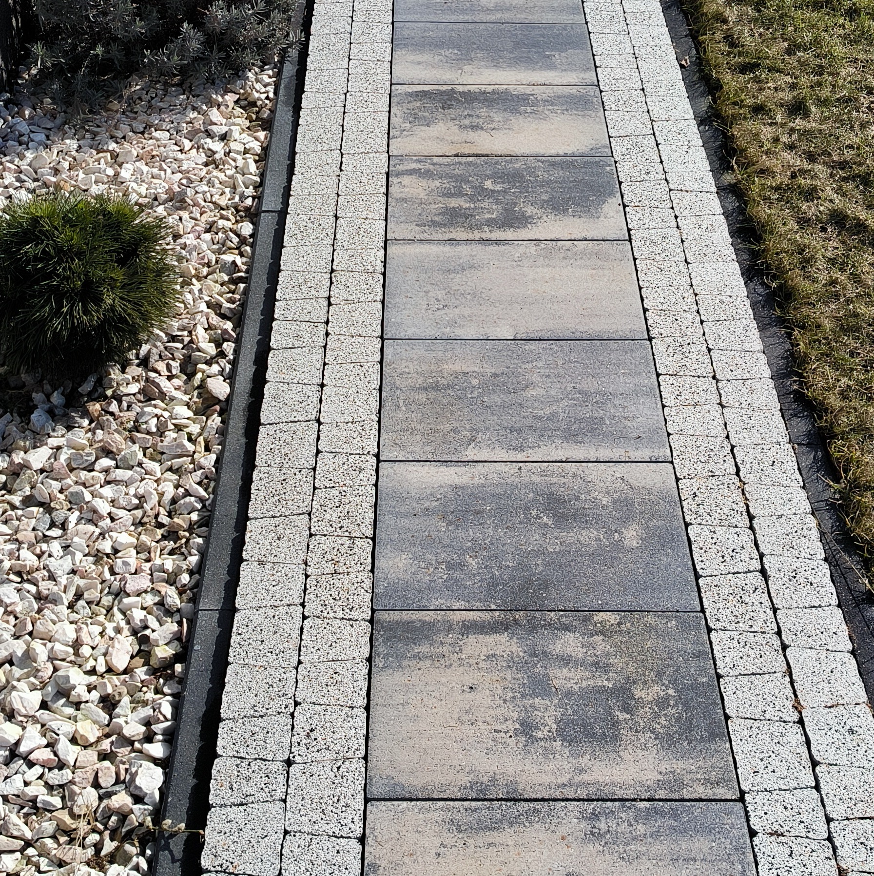 Ścieżka z szarej kostki brukowej, obrzeża z jasnej kostki, po bokach białe kamienie i trawa.
