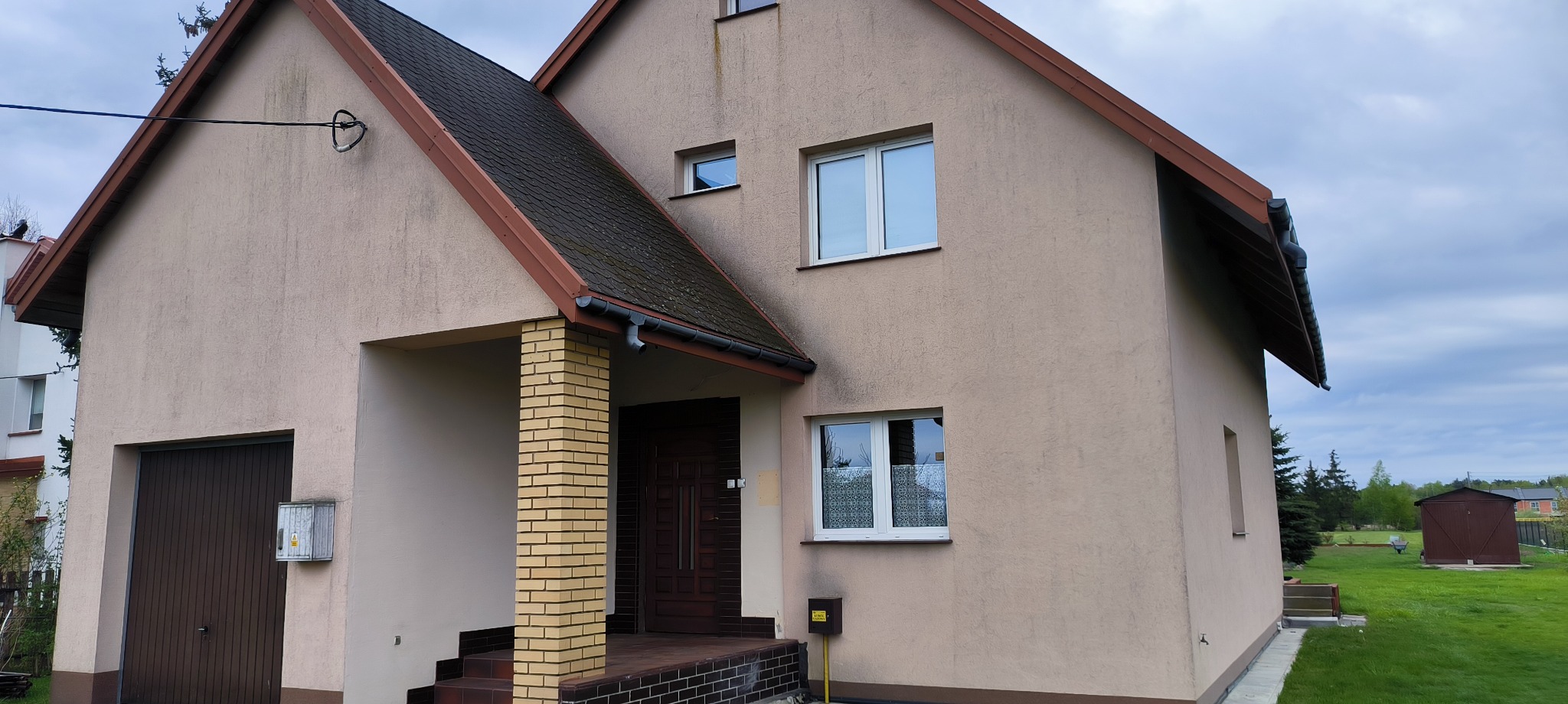 Dwukondygnacyjny dom z beżową elewacją i ciemnym dachem. Widoczne ślady zabrudzeń na elewacji, szczególnie pod oknami. Wejście z kolumną z żółtej cegły.