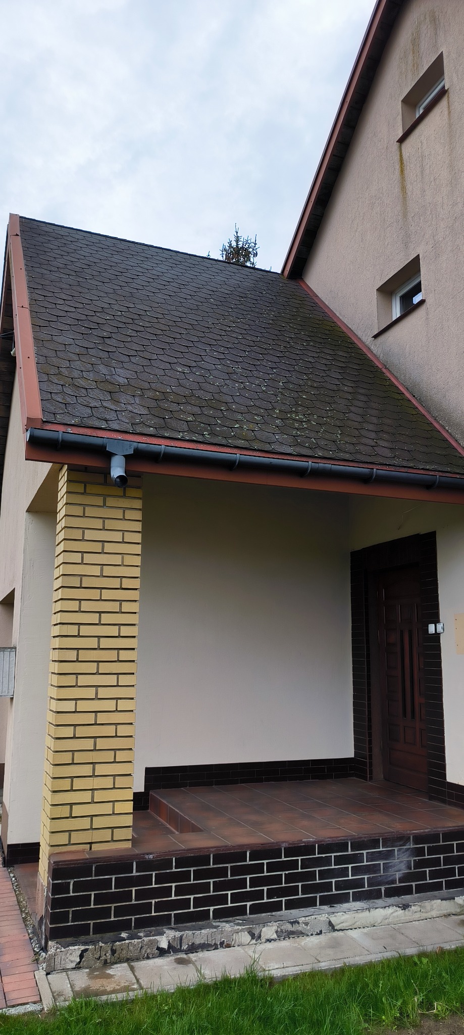 Dom z zadaszonym wejściem, widoczny dach pokryty bitumiczną dachówką z mchem, ceglana kolumna i fragment elewacji. Widoczne ślady zabrudzeń na dachu.