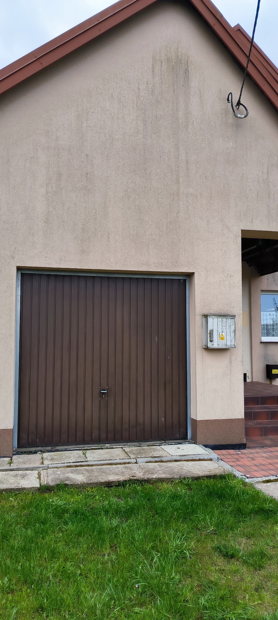 Brudna elewacja domu z brązową bramą garażową i skrzynką elektryczną. Widoczne ślady zacieków na elewacji. Fragment trawnika przed budynkiem.