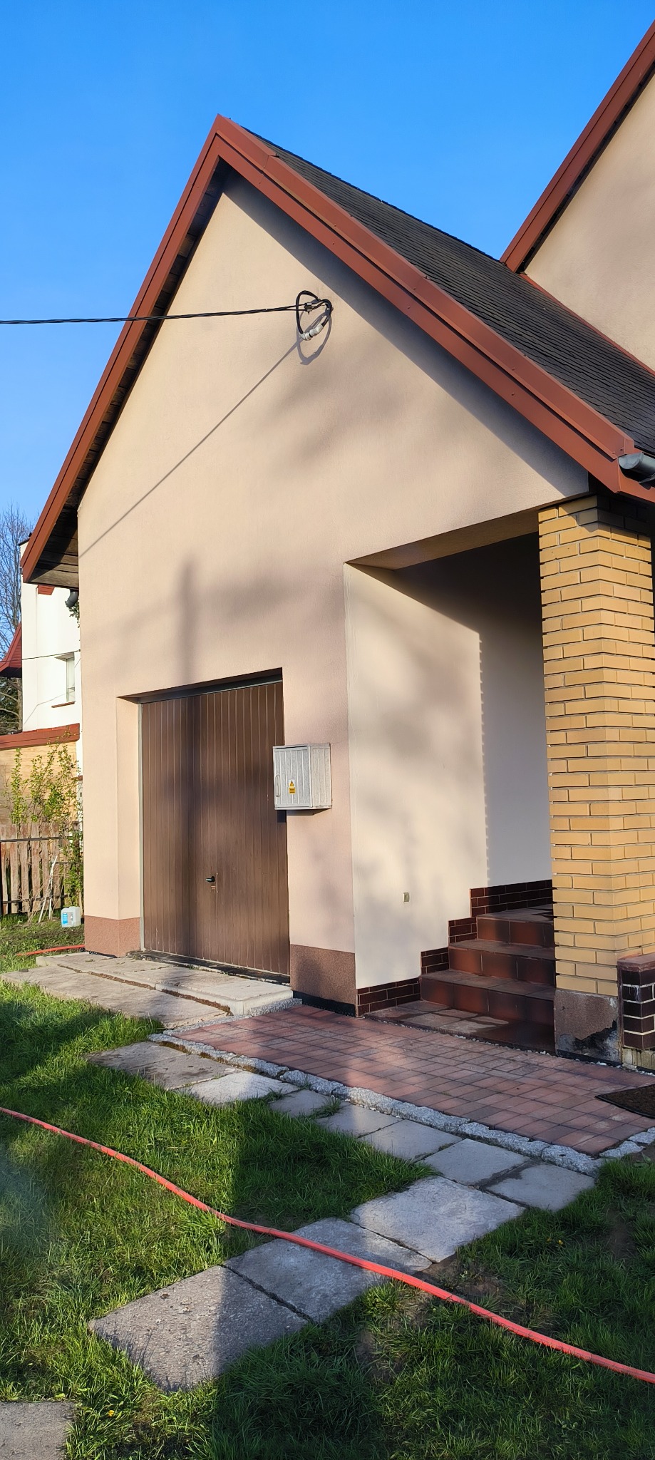 Dom z beżową elewacją, brązowym dachem i garażem. Widoczne schody z cegły klinkierowej i trawnik. Elementy instalacji na elewacji. Słoneczny dzień, błękitne niebo.