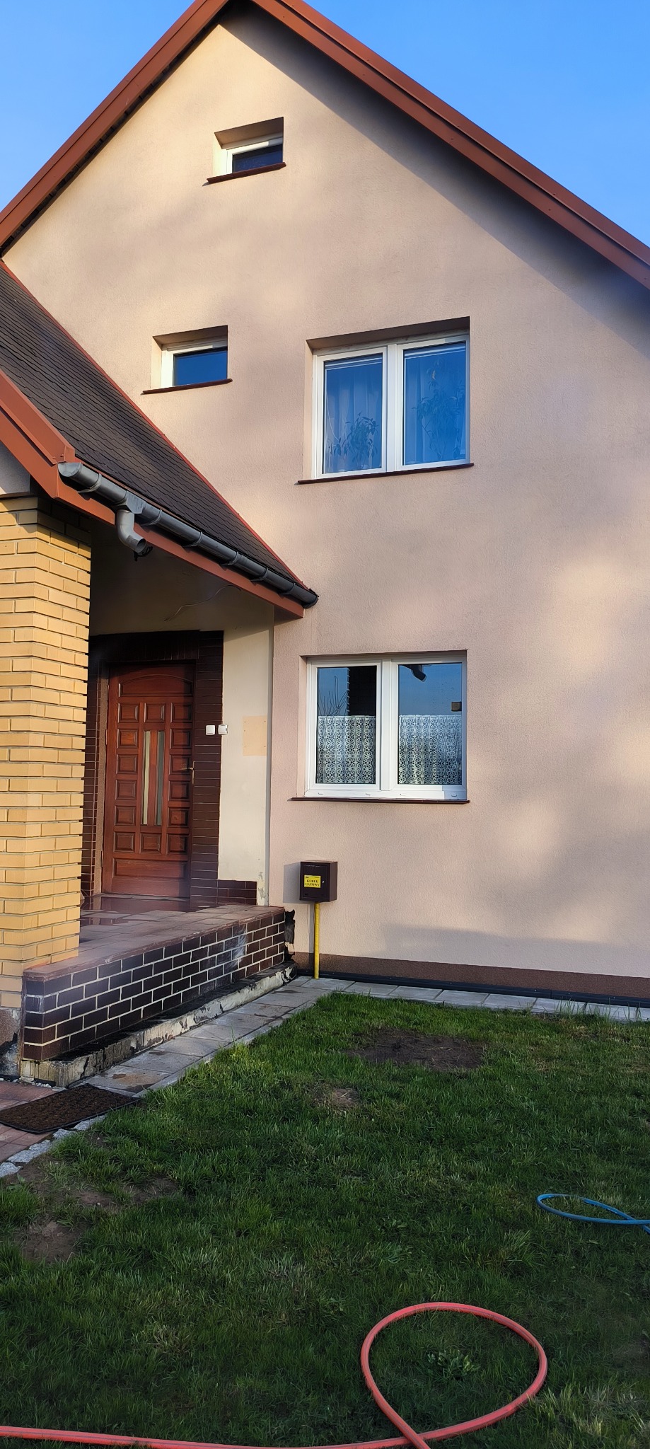 Jasna elewacja dwupiętrowego domu z brązowym dachem i drewnianymi drzwiami wejściowymi. Widoczne dwa okna na piętrze i jedno na parterze.