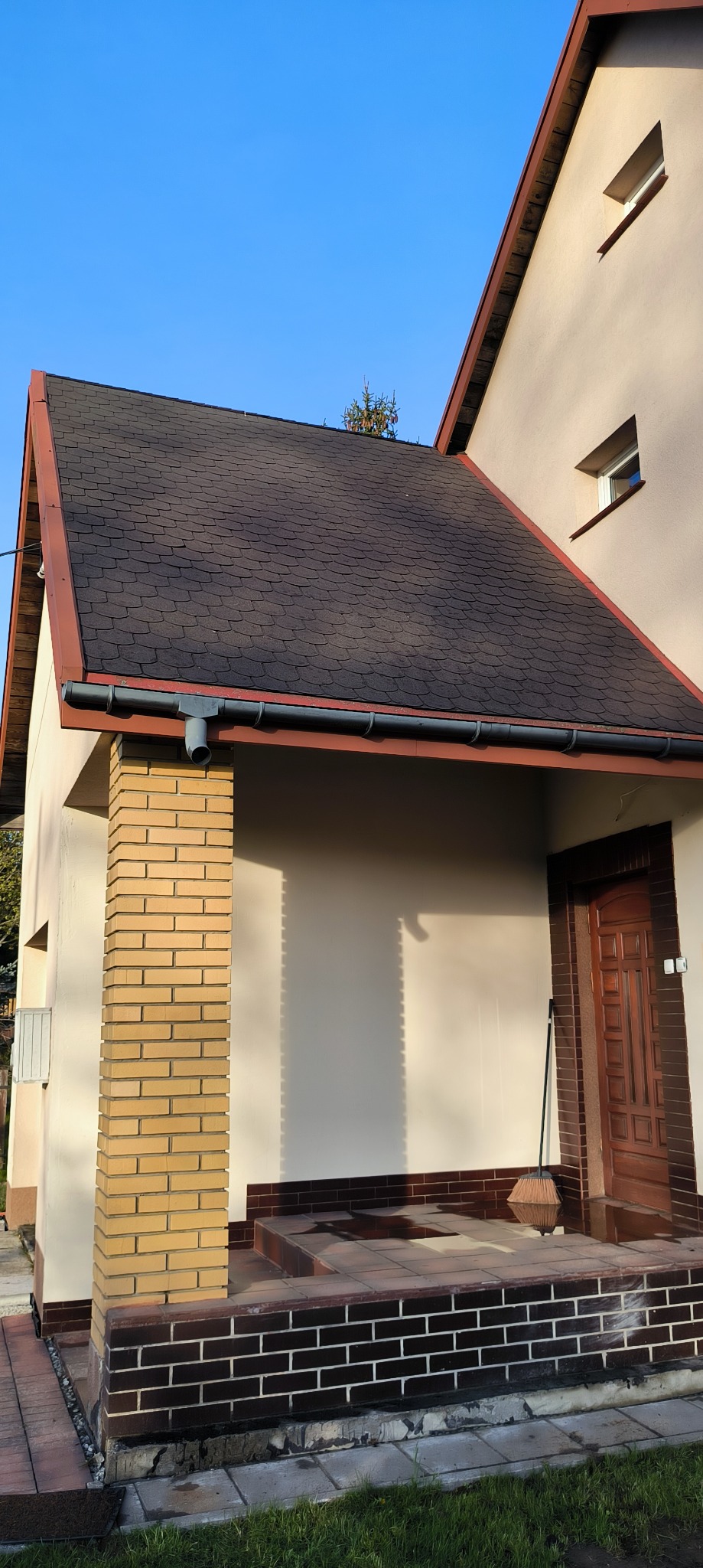 Dom z dachem pokrytym gontem bitumicznym w odcieniach brązu. Widoczna elewacja w jasnym kolorze, ceglana kolumna i drewniane drzwi wejściowe. Na dachu rośnie małe drzewko.