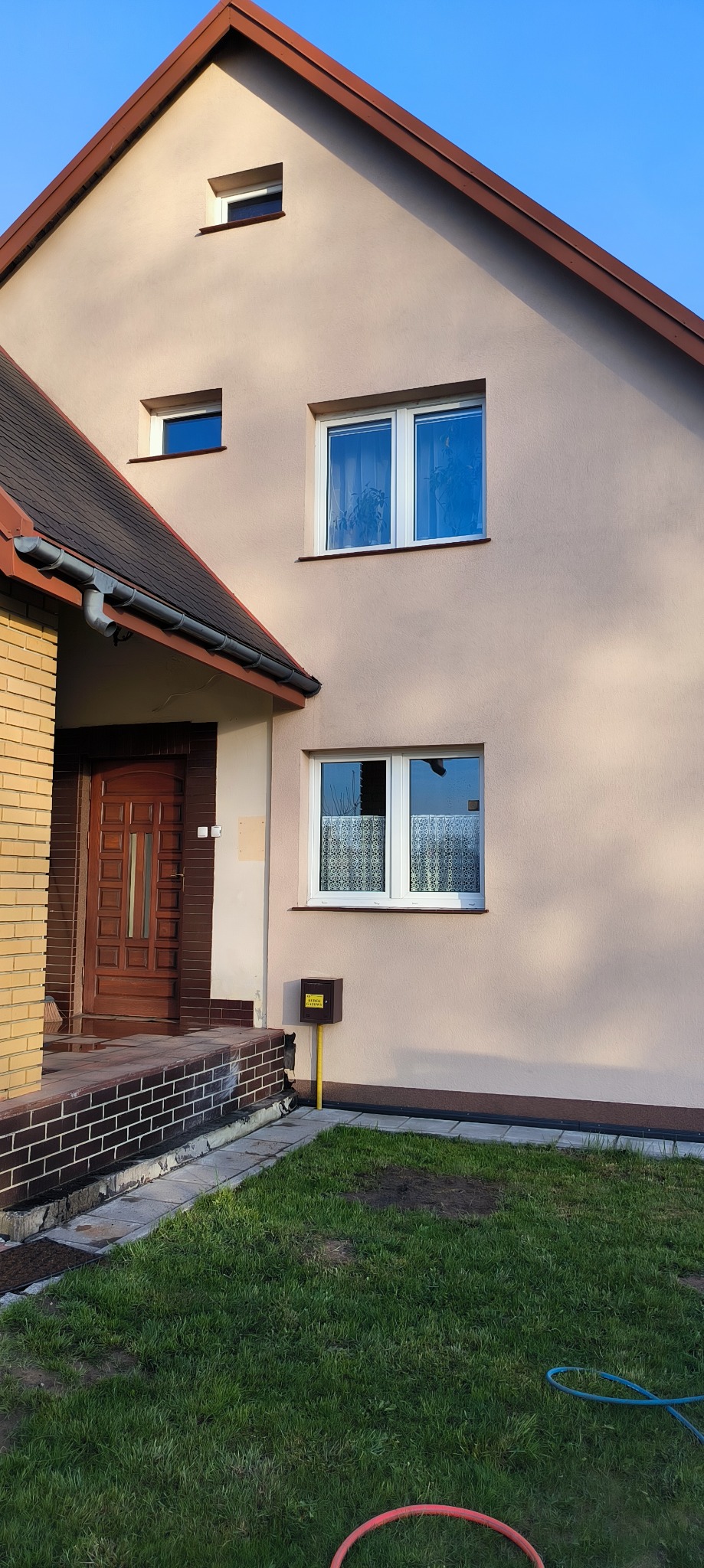 Jasna elewacja domu jednorodzinnego z brązowym dachem i stolarką okienną, widok z poziomu trawnika, skrzynka gazowa przy ścianie.