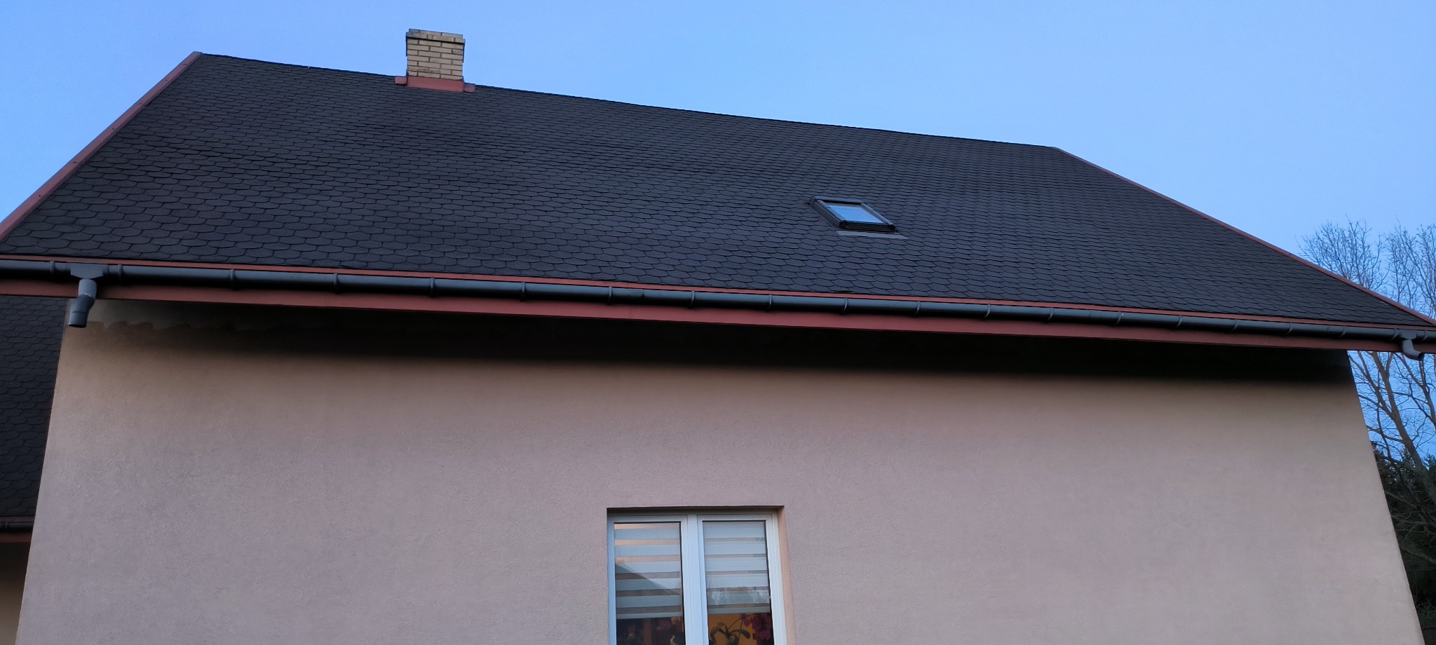 Ciemny dach z gontu bitumicznego z oknem dachowym i ceglanym kominem na tle błękitnego nieba. Elewacja budynku w kolorze beżowym z oknem.