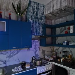 Rafi-Bud - Niebieska kuchnia z marmurowymi płytkami, na blacie kuchenka z garnkiem, widoczne szafki górne z dekoracjami i trofeum.