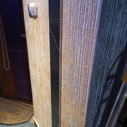 Rafi-Bud - Fragment ściany z dekoracyjną fakturą w odcieniach beżu i szarości, z pionowym pasem czarnego kamienia i włącznikiem światła. Widoczny fragment wycieraczki pod drzwiami.