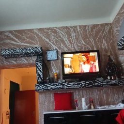Rafi-Bud - Ściana w salonie z efektem marmuru, dekoracyjna półka pod telewizorem w czarno-białe wzory, widoczny fragment futryny drzwi i zegar na ścianie.