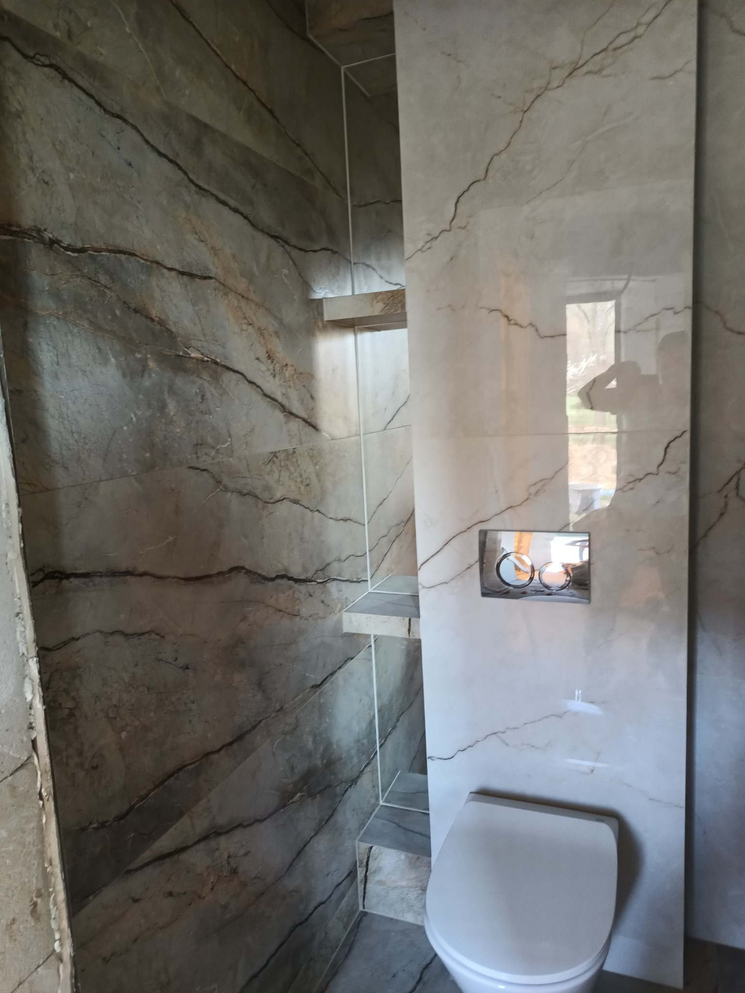 Nowoczesna łazienka z marmurowymi płytkami w odcieniach szarości i bieli, podwieszana toaleta z chromowanym przyciskiem spłukującym, szklane półki w rogu kabiny prysznicowej.