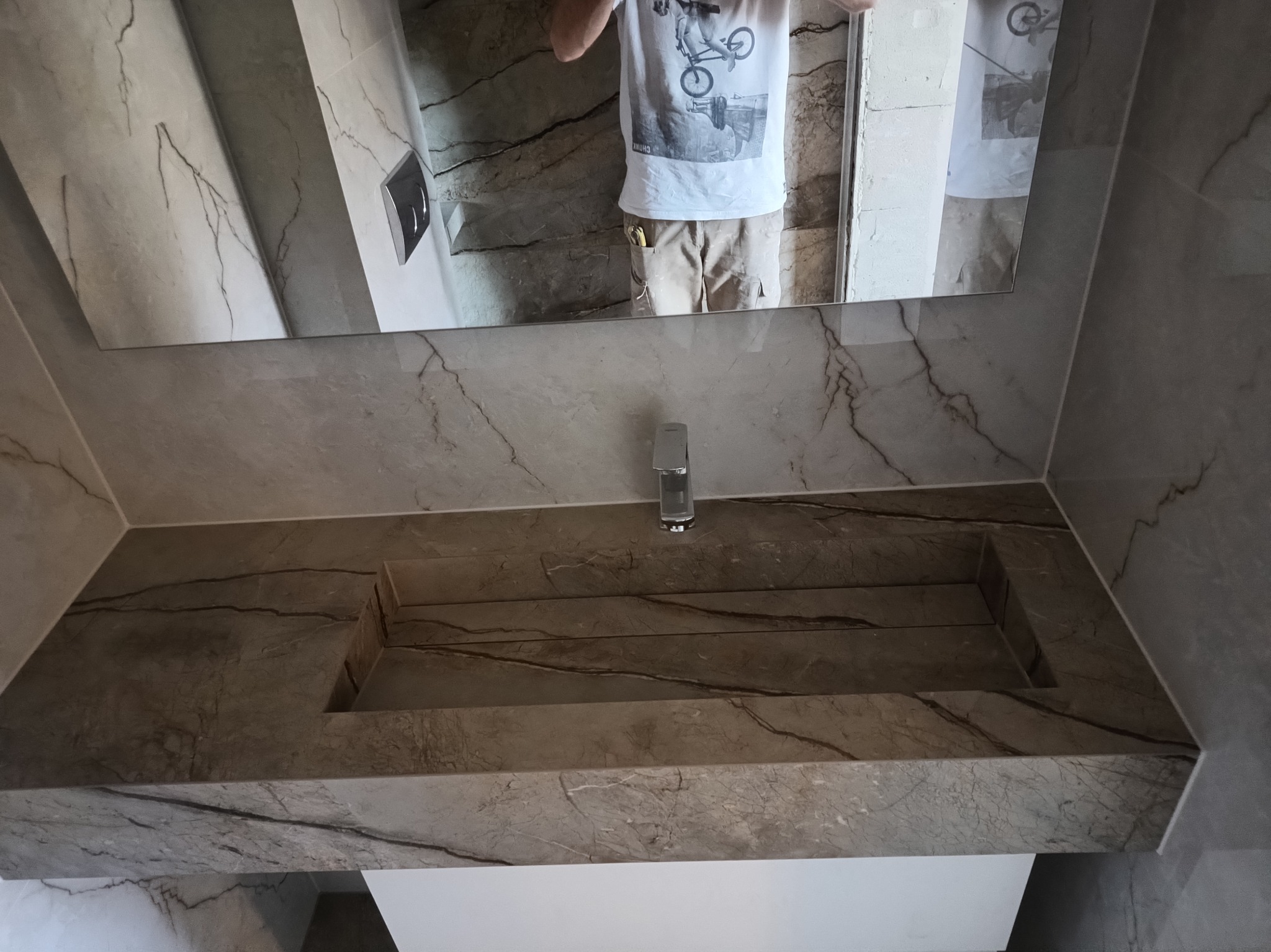 Łazienka z umywalką wpuszczoną w blat z szarego marmuru z widocznym odbiciem osoby w lustrze.