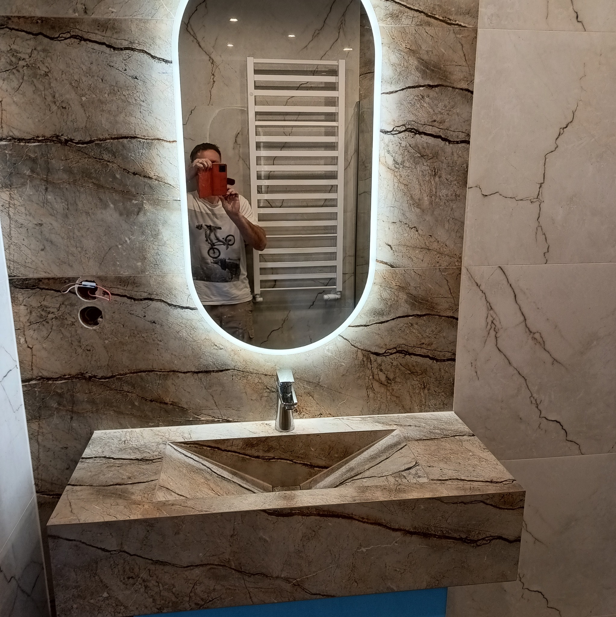Umywalka z kamienia o unikalnym wzorze w łazience z marmurowymi płytkami, widoczne oświetlone lustro owalne i grzejnik drabinkowy, odbicie fotografa z telefonem.