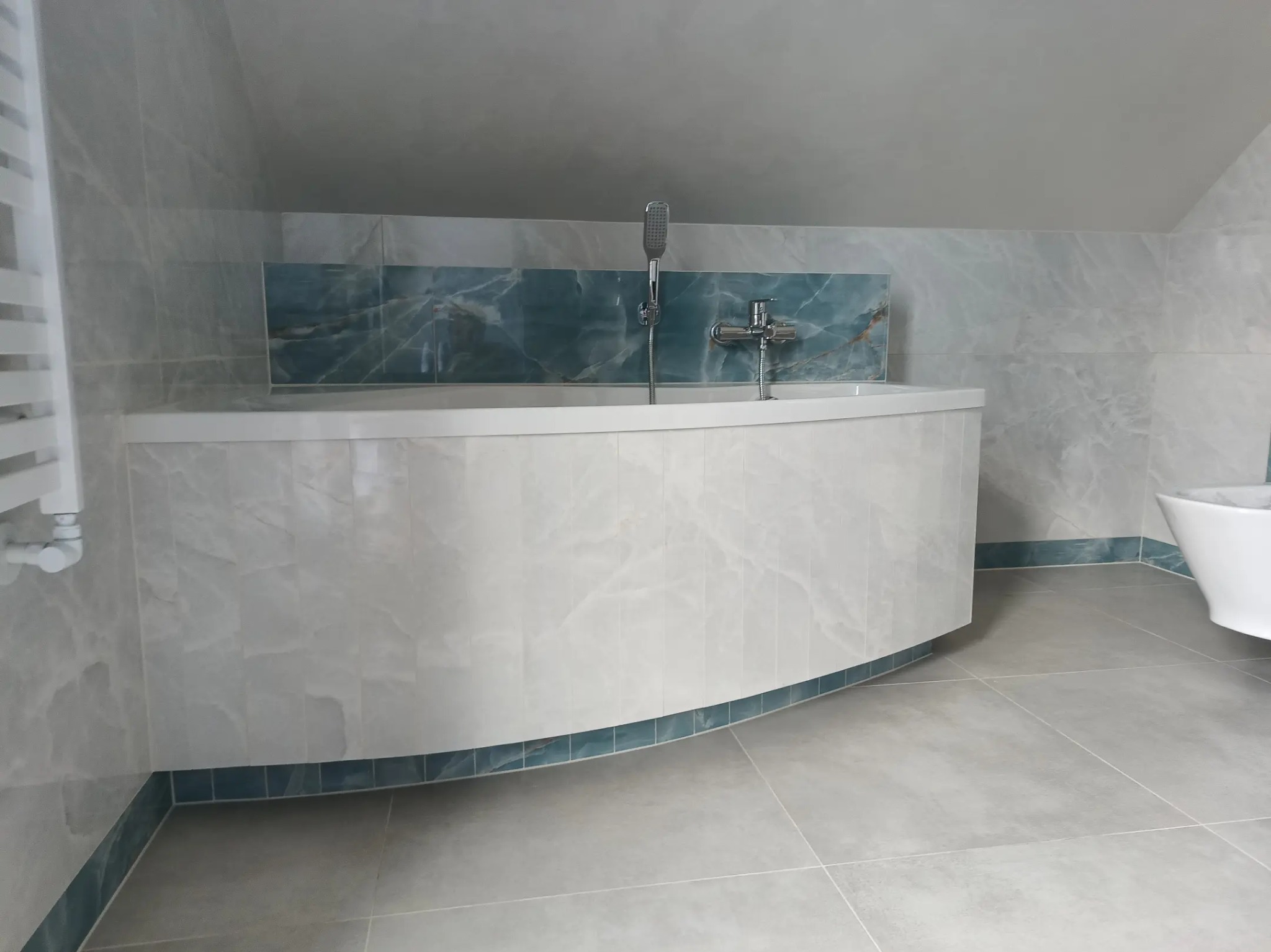 Zaokrąglona wanna obudowana płytkami imitującymi marmur, z niebieskim akcentem w postaci płytek dekoracyjnych nad wanną i przy podłodze, w łazience na poddaszu ze skośnym sufitem.