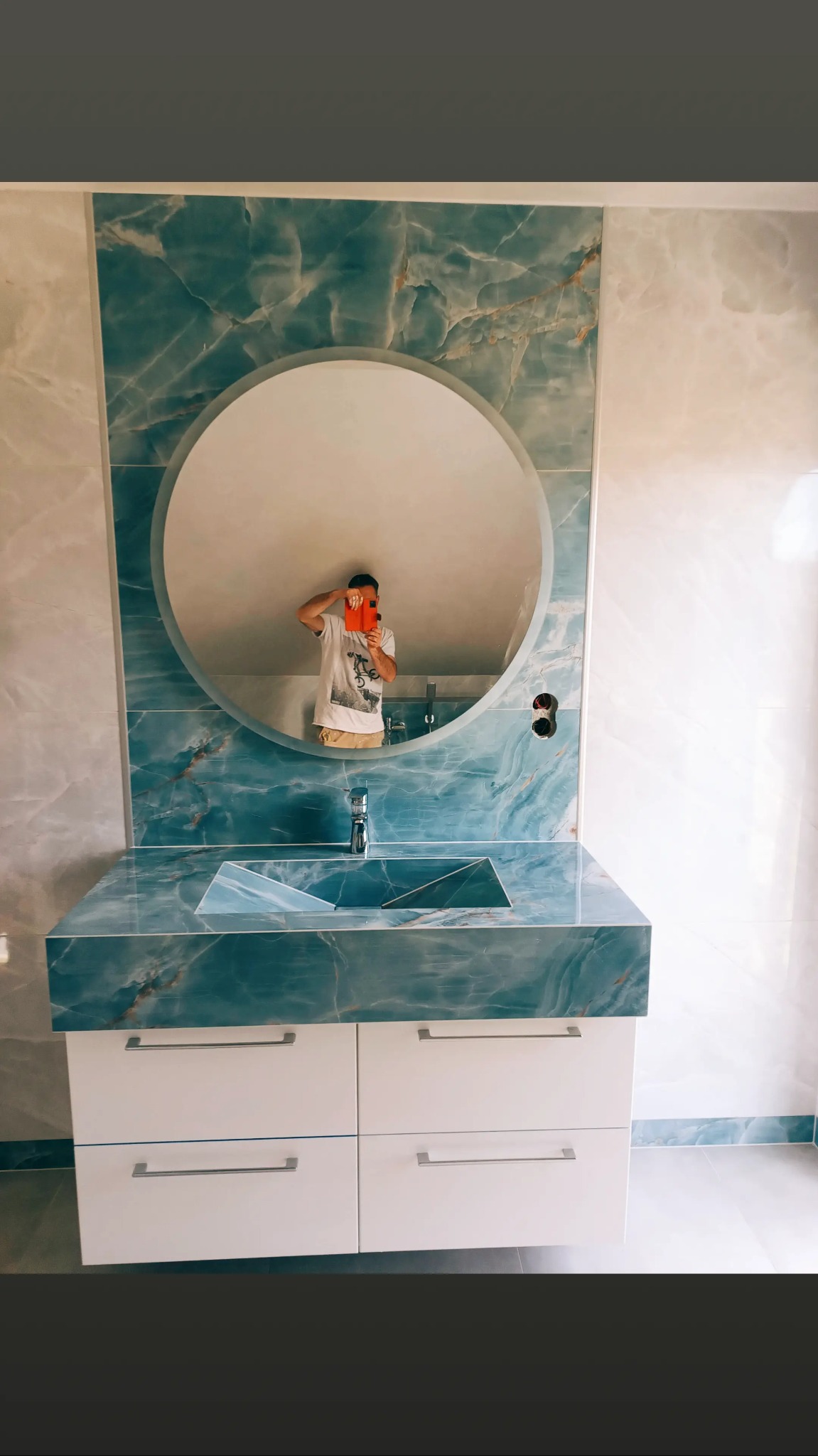 Nowoczesna łazienka z umywalką nablatową z niebieskiego marmuru, okrągłe lustro z odbiciem fotografa, białe szafki pod umywalką i marmurowe płytki na ścianach.