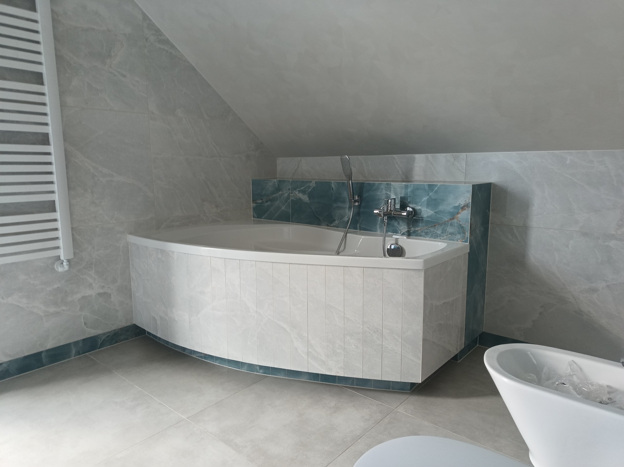 Wykończona łazienka na poddaszu z wanną narożną obudowaną panelami imitującymi marmur, szarymi płytkami ściennymi i podłogowymi oraz grzejnikiem drabinkowym po lewej stronie.