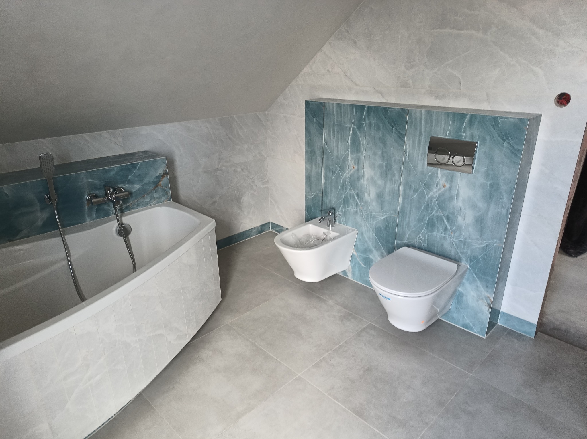 Nowoczesna łazienka na poddaszu z marmurowymi płytkami w odcieniach szarości i błękitu, z wanną narożną, bidetem i toaletą podwieszaną z chromowanym przyciskiem spłukującym.