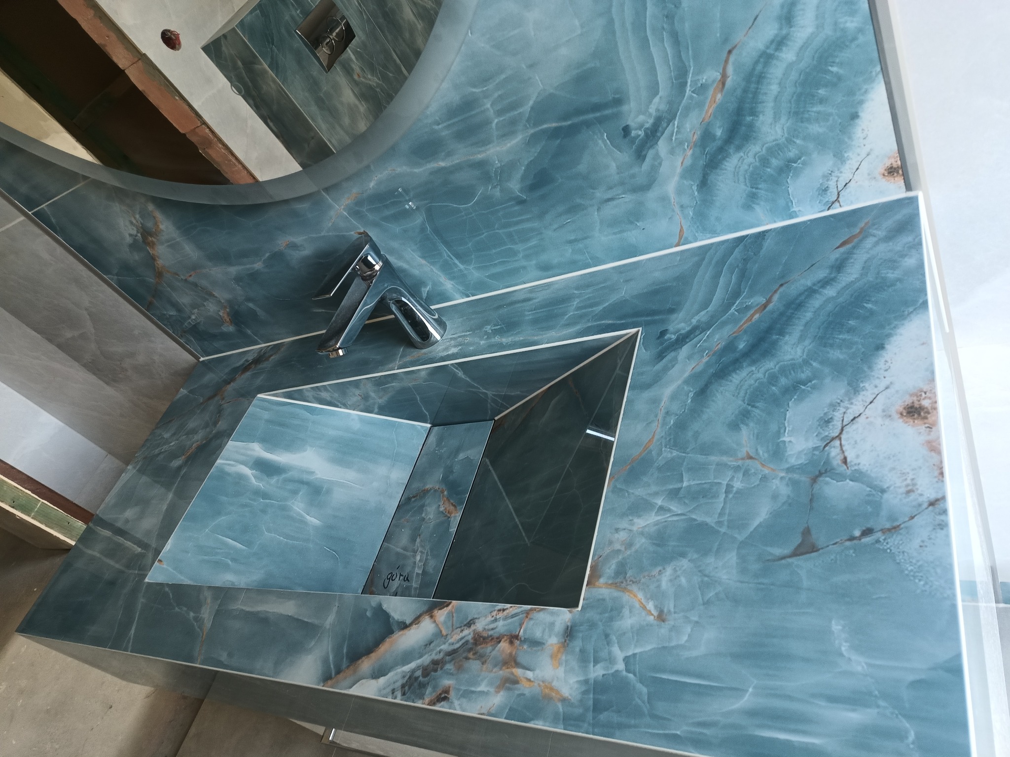 Wykończona łazienka z umywalką wpuszczoną w blat z płytek imitujących marmur w odcieniach błękitu i szarości, nowoczesna armatura, widoczne fragmenty surowego sufitu i ściany w trakcie remontu.