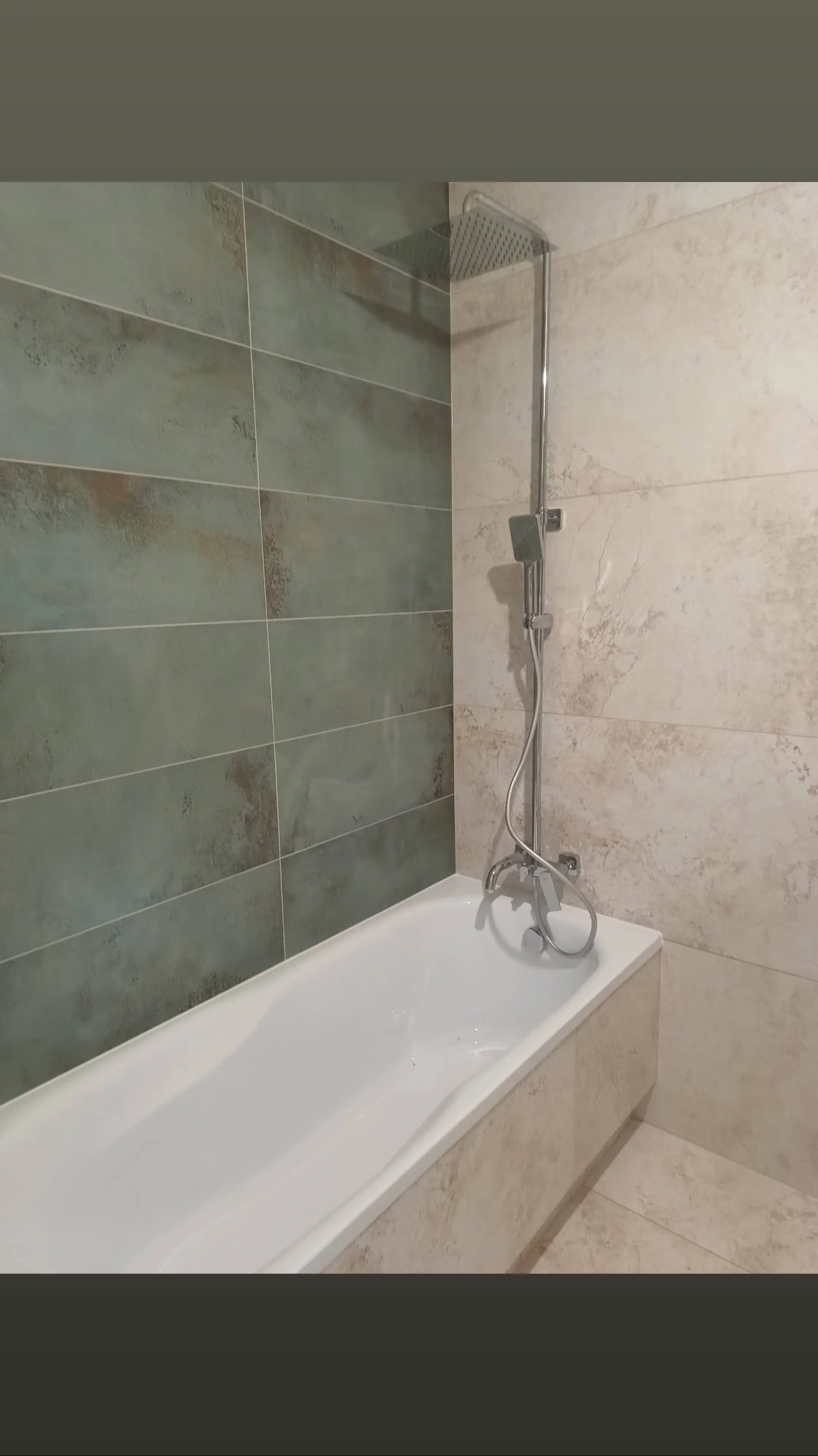 Nowoczesna łazienka z wanną obudowaną beżowymi płytkami imitującymi marmur, ściany wyłożone płytkami w odcieniach zieleni i beżu, bateria wannowa z deszczownicą.