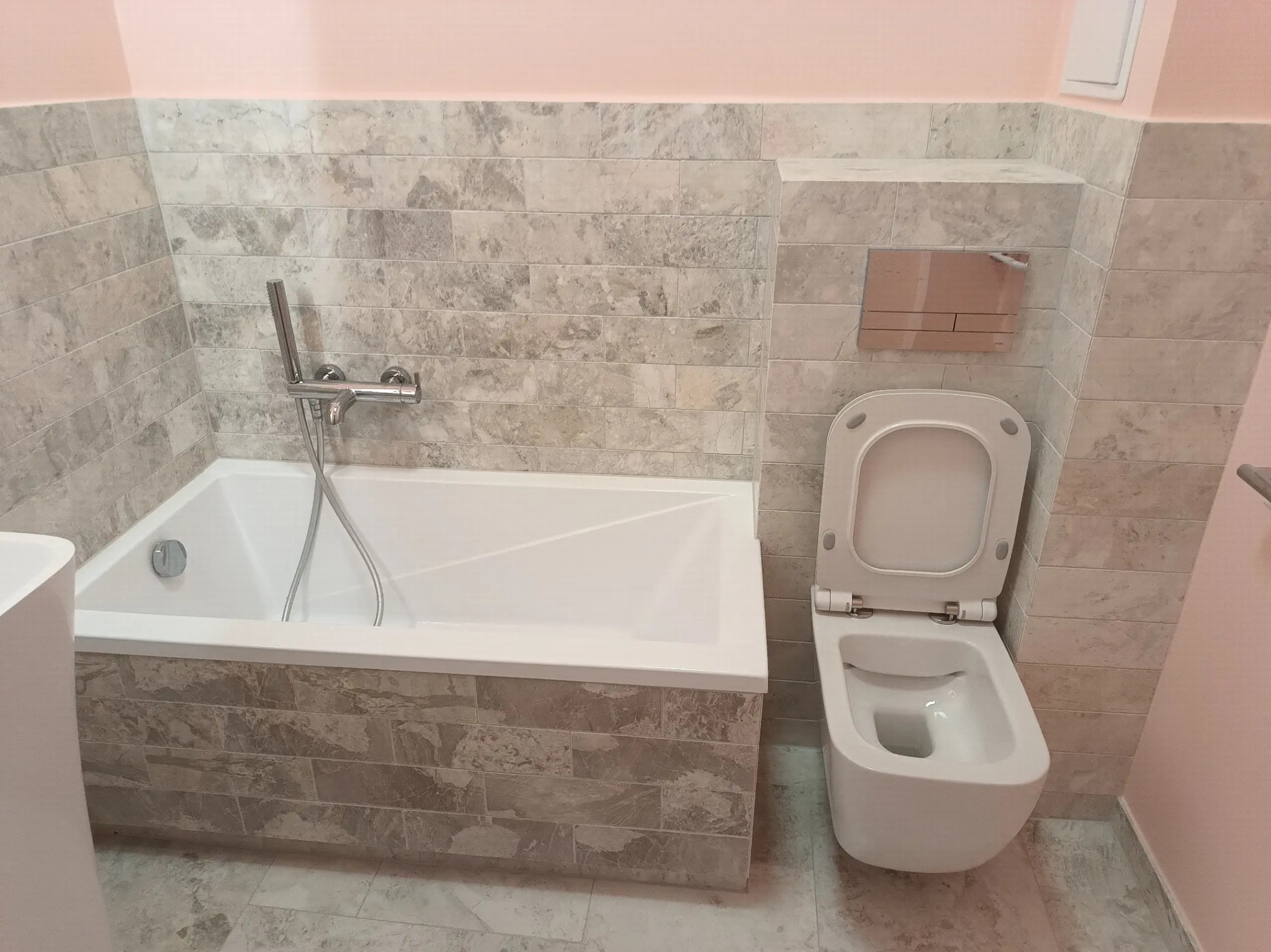 Wykończona łazienka z wanną obudowaną płytkami imitującymi kamień i podwieszaną toaletą z otwartą klapą, ściany wyłożone podobnymi płytkami, różowa ściana w rogu.
