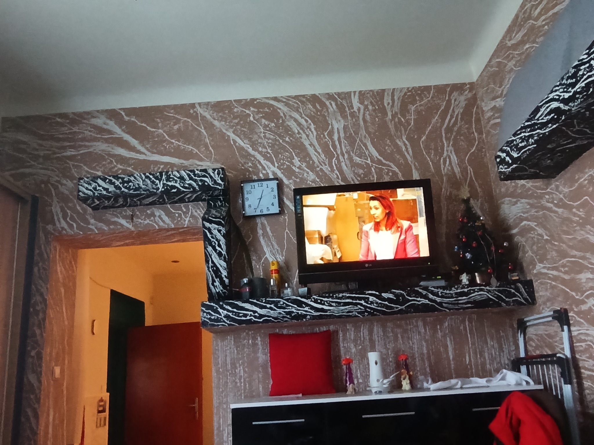 Ściana w salonie z efektem marmuru, dekoracyjna półka pod telewizorem w czarno-białe wzory, widoczny fragment futryny drzwi i zegar na ścianie.