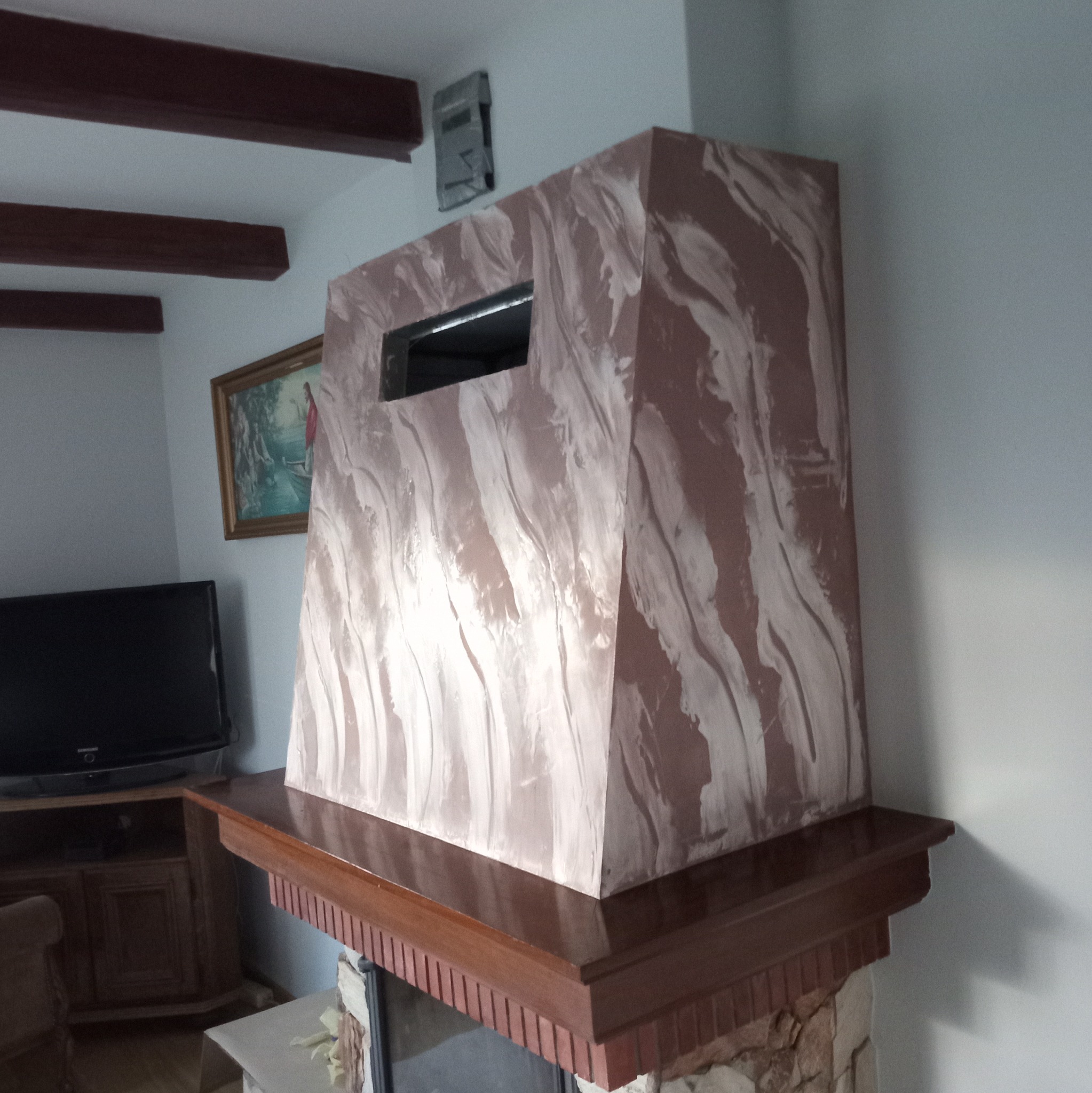 Dekoracyjna obudowa kominka z efektem marmuru w odcieniach brązu i beżu, z prostokątnym otworem, umieszczona na drewnianym gzymsie kominkowym z ceglanym wykończeniem, w tle telewizor i obraz.