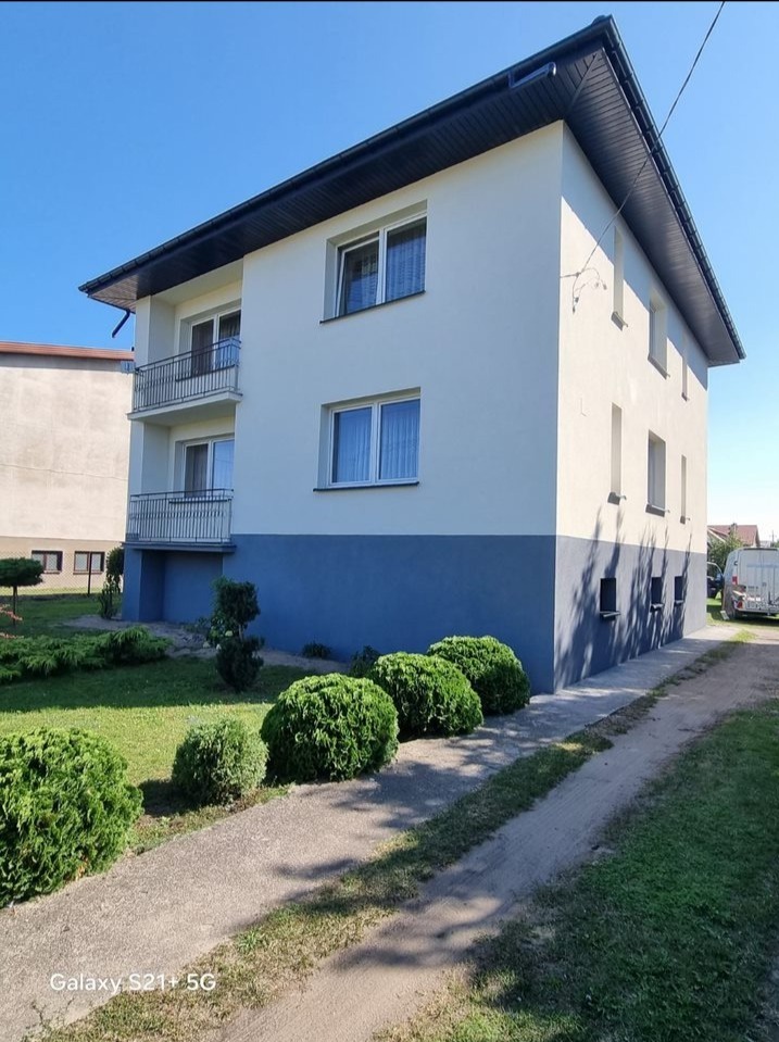 Dwukondygnacyjny budynek z balkonami i dwukolorową elewacją (kremowa góra, niebieski dół), widok z poziomu trawnika z ozdobnymi krzewami.