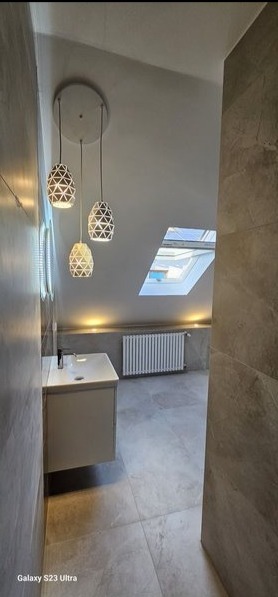 Nowoczesna łazienka na poddaszu z marmurowymi płytkami, wiszącą szafką z umywalką, trzema lampami wiszącymi o geometrycznym wzorze i oknem dachowym w skosie.