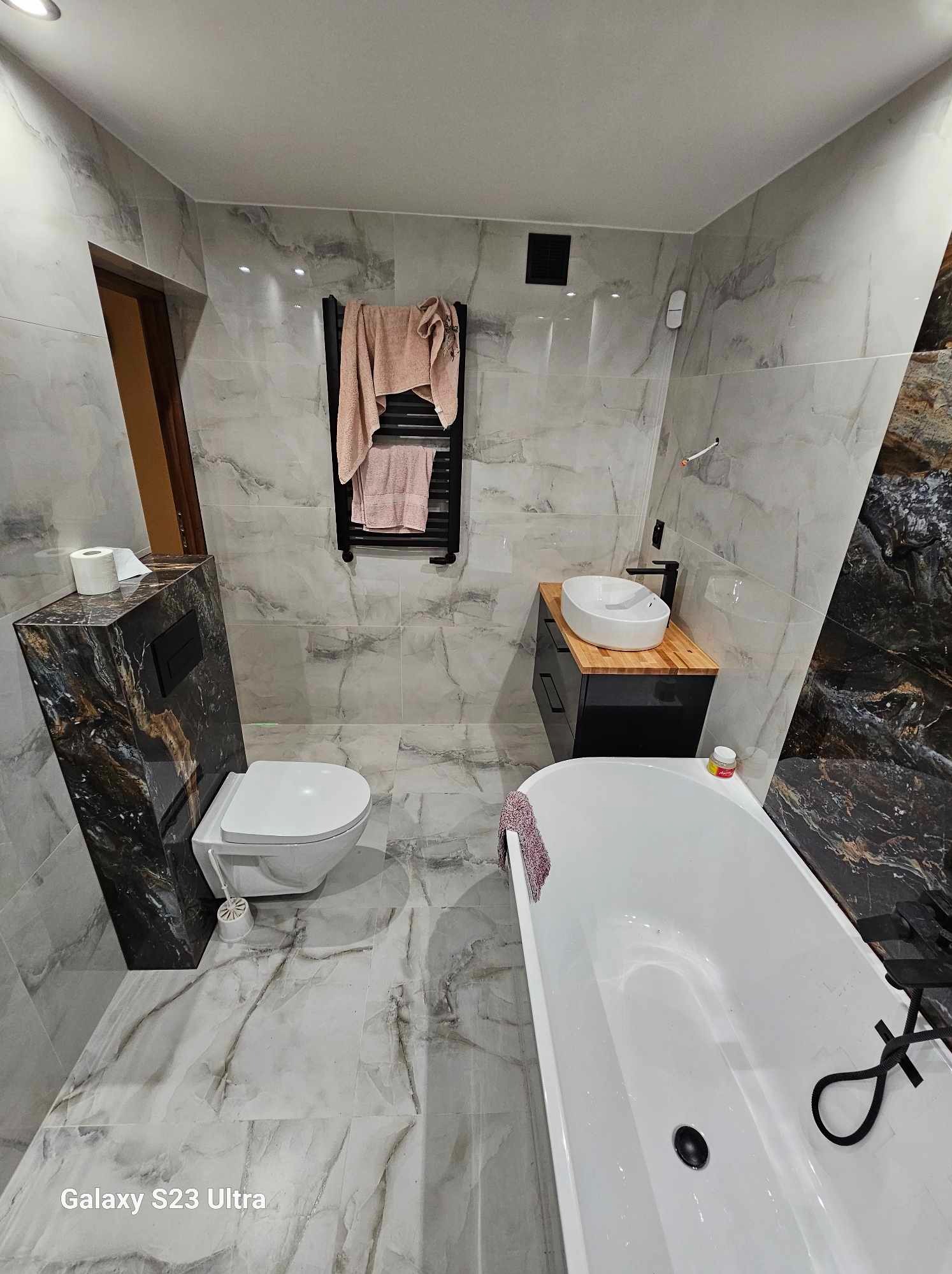 Nowoczesna łazienka z marmurowymi płytkami, wiszącą toaletą z czarnym panelem, białą umywalką na drewnianym blacie i białą wanną z czarną armaturą.