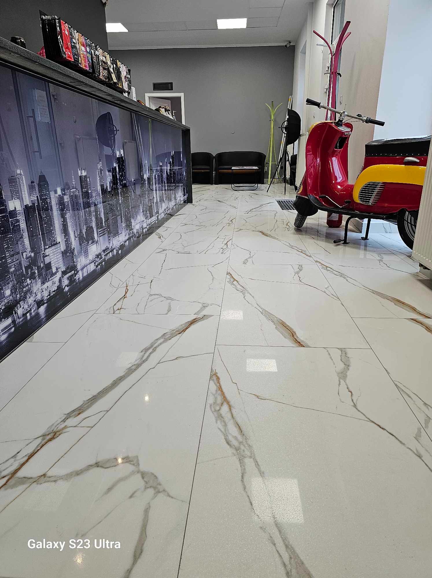 Białe marmurowe płytki podłogowe z brązowymi żyłami, widok na fragment czerwono-żółtego skutera, czarna lada z grafiką panoramy miasta i meble w tle.