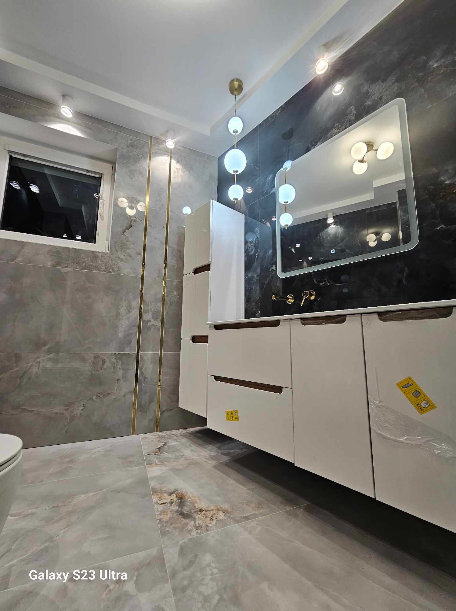 Nowoczesna łazienka z szarymi marmurkowymi płytkami, białymi szafkami pod umywalkę z drewnianymi akcentami, podświetlanym lustrem i dekoracyjnymi lampami wiszącymi.