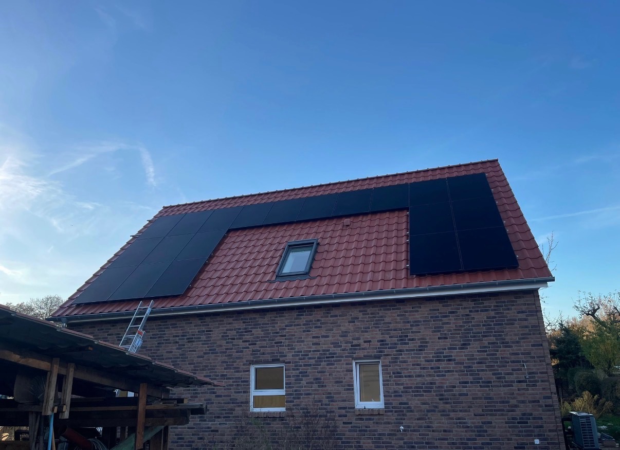 Częściowy widok domu z cegły z panelami fotowoltaicznymi na czerwonym dachu. Na dachu widoczne okno dachowe. Z boku domu oparta drabina.