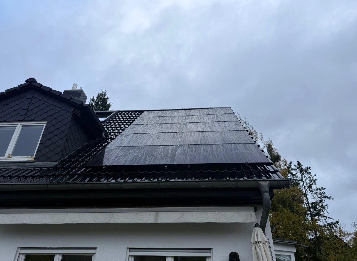 Czarne panele słoneczne na dachu domu w deszczowy dzień. Widoczne dachówki, okno poddaszowe i fragment elewacji. Pochmurne niebo w tle.