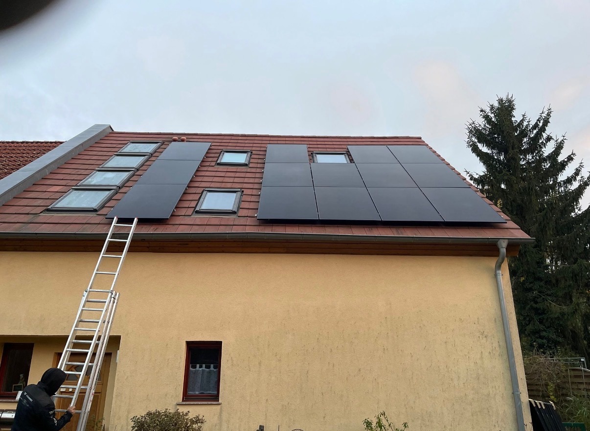 Montaż paneli na dachu z oknami połaciowymi. Aluminiowa drabina oparta o ścianę budynku. Widok na zamontowane panele w pochmurny dzień.