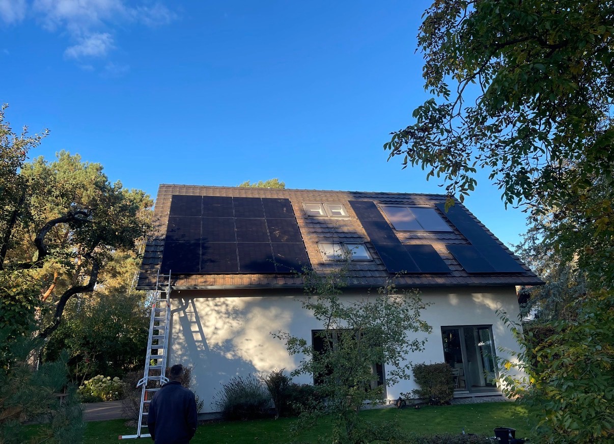 Dom jednorodzinny z panelami słonecznymi na dachu, drabina oparta o ścianę, widok z dołu, błękitne niebo, drzewa w tle.