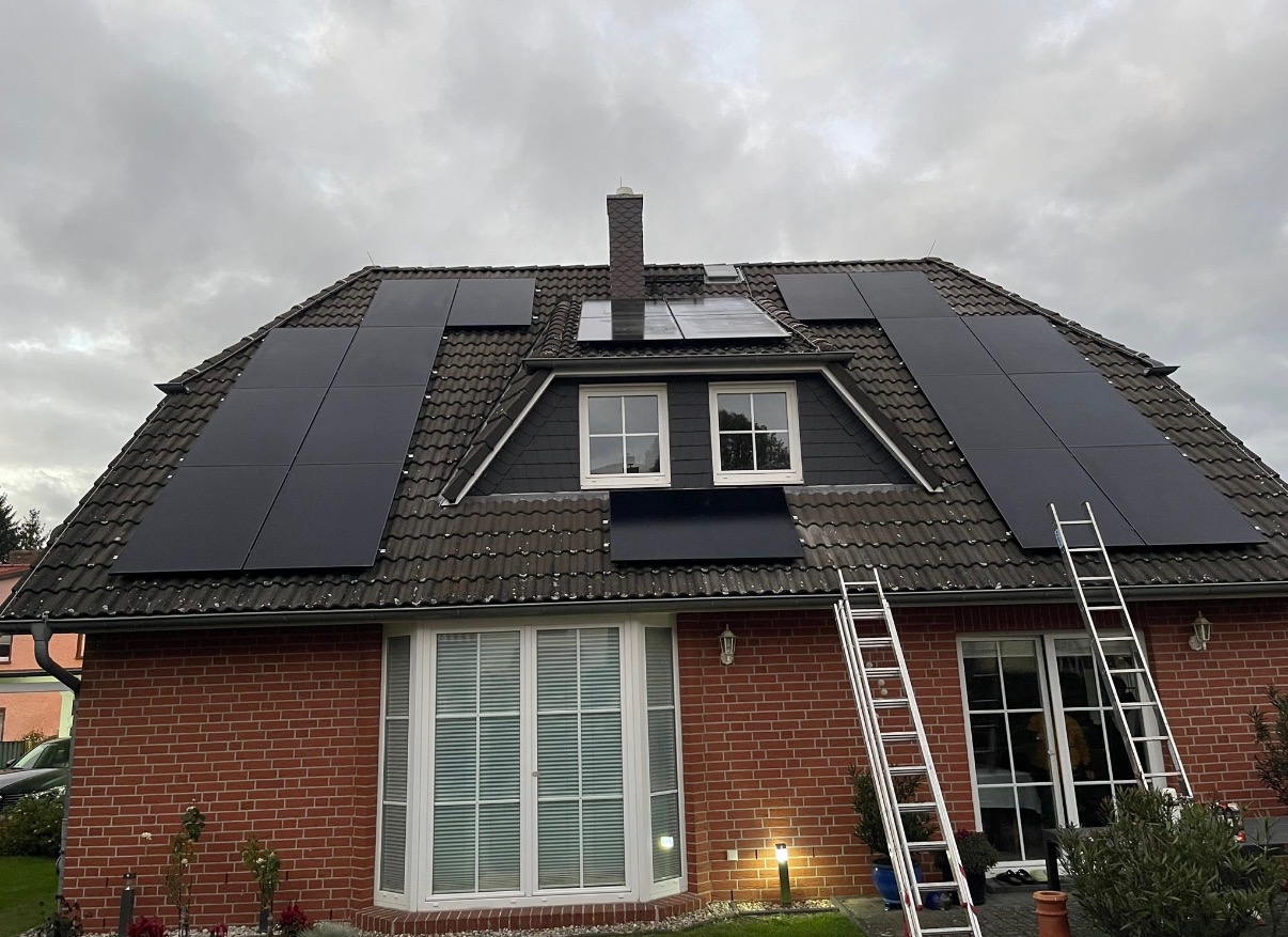 Dom z czerwonej cegły z panelami słonecznymi na dachu, drabiny opierają się o budynek. Pochmurne niebo w tle. Widok z dołu.